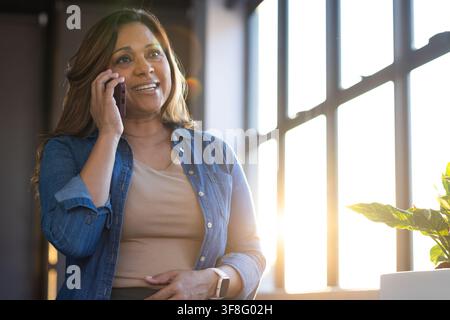 Reife afrikanisch-amerikanische Frau telefoniert am Smartphone nahe einem Fenster, Jeanshemd und Smartwatch sichtbar Stockfoto