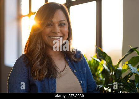 Afroamerikanische Frau steht an einem geteilten Fenster, Sonne scheint, trägt Denimhemd, beiges Oberteil, Pflanzen Stockfoto