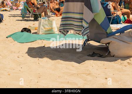 Nahaufnahme von Strandaccessoires aus Sand, darunter gestreifte Handtücher, eine Tragetasche mit Text, Flip-Flops und Strandliegen mit Menschen, die in der Sonne liegen Stockfoto