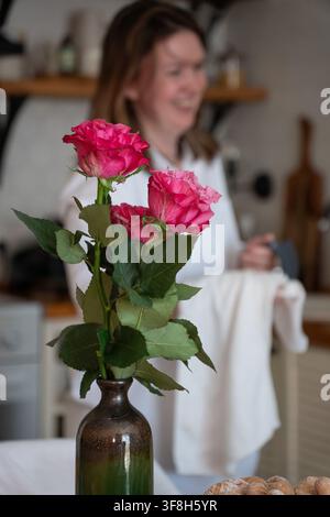 Rosa Rosen in der Vase stehen im Fokus, während eine lächelnde Frau in der Küchenwohnung im verschwommenen Hintergrund arbeitet. Blumen, Wohnstil, häusliches Leben, Blumendekor Stockfoto