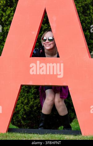 FRAU SCHAUT DURCH EINEN GIGANTISCHEN ROTEN BUCHSTABEN A, REPORTAGE, GREEN MAN FESTIVAL 2016, BRECON, WALES, UK, 2016: Eine lächelnde junge Frau mit blondem Haar und runden reflektierenden Sonnenbrillen schaut verspielt durch die dreieckige Öffnung eines großen, freistehenden roten Buchstabens „A“ im Freien. Foto: Rob Watkins. INFO: Das Green Man Festival begann 2003 in Wales. Jährlich findet es am dritten Wochenende im August in den Brecon Beacons statt und feiert alternative Musik, Kunst, Literatur, Wissenschaft und familienfreundliche Kultur in einer wunderschönen Berglandschaft. Stockfoto