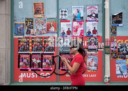Junge Frau schaut auf ihr Handy und geht an Edinburgh-Fringe-Show-Plakaten am Fenster eines leeren Ladens vorbei. Stockfoto