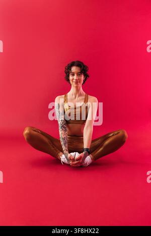 Junge schlanke Frau in goldener Fitnessbekleidung, die auf einem purpurroten Hintergrund sitzt Stockfoto