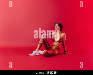 Junge lächelnde Frau in goldener Fitnessbekleidung, die auf einem purpurroten Hintergrund sitzt und nach oben schaut Junge lächelnde Frau in goldener Fitnessbekleidung, die auf einem Stockfoto