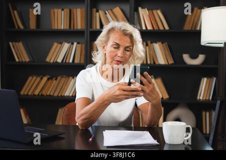 Selbstbewusste, stilvolle europäische Seniorin mittleren Alters mit Smartphone am Arbeitsplatz. Stilvolle ältere Geschäftsfrau aus den 60er Jahren mit Handy im Büro. Boss Leader mit Internet-Apps Stockfoto