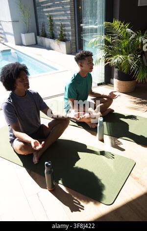 Afroamerikanische Männer, die im Schneidersitz auf grünen Yogamatten neben einer Schiebetür mit Flaschen sitzen Stockfoto