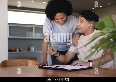 Verschiedene männliche Freunde lachen, lehnen sich an einen hölzernen Esstisch in der Küche und überprüfen einen Ordner mit einem Stift Stockfoto