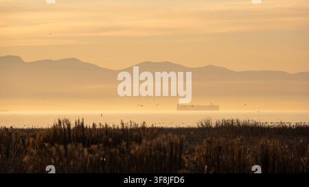 Sonnenuntergang an einem Gewässer, wo das Sonnenlicht durchbricht und Berge sowie ein im Nebel schwebendes Boot beleuchtet. Vögel fliegen darüber und sorgen für eine lebhafte Szenerie Stockfoto