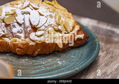 Ansicht eines köstlichen Mandelcroissants mit geschnittenen Mandeln und einem Hauch Puderzucker auf einem strukturierten blaugrünen Teller in London, England, Vereinigtes Königreich Stockfoto