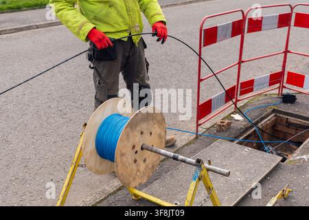 Arbeiter in einer gelb-grünen Warnjacke und roten Handschuhen legt schwarzes Glasfaserkabel von Hand, mit einer blauen Seiltrommel, die auf einem gelben Kabel montiert ist Stockfoto