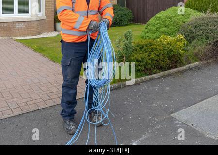 Arbeiter in einer orangefarbenen Warnjacke und dunklen Hosen sammelt eine große Spule blauen Zugseils von Hand auf einer Wohnhausauffahrt. Schmutzige Handschuhe an Stockfoto