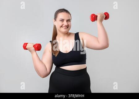 Glückliche übergewichtige Frau mit Hanteln auf hellgrauem Hintergrund Stockfoto