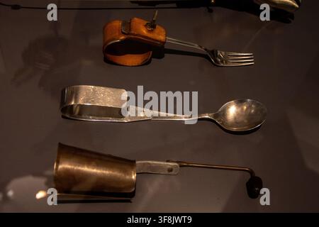 WIEN, ÖSTERREICH, 7. Februar 2023: Drei alte adaptive Utensilien, darunter eine Gabel, ein Löffel und ein Griffbecher, werden hinter Glas in einer Museumsausstellung präsentiert Stockfoto