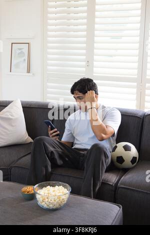 Asiatischer Mann Anfang 20 auf dunklem grauen Sofa sitzend, der ein Smartphone hält, sein Haar berührt, Fußballball in der Nähe Stockfoto