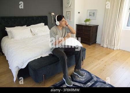 Asiatischer Mann trägt graues T-Shirt, passt weiße Kopfhörer an, während er im Schlafzimmer auf einer gepolsterten Bank singt Stockfoto
