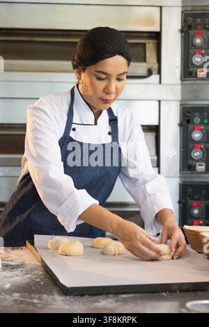 Bäckerin formt Teigkugeln auf einem Tablett in einer Bäckerei mit Öfen, trägt Schürzenjacke Stockfoto