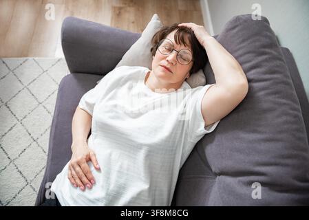 Kranke ältere Frau, die an saisonaler Grippe oder Erkältung leidet und auf dem Sofa liegt. Kranke Seniorin fühlt sich mit Influenza zu Hause unwohl Stockfoto