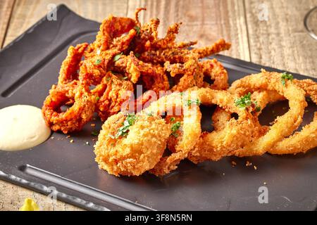 Knuspriges frittiertes Hühnerpopcorn und Calamariringe, serviert in einem Korb und auf einem schwarzen Tablett mit Dipsauce auf einem rustikalen Holzuntergrund, Restaura Stockfoto