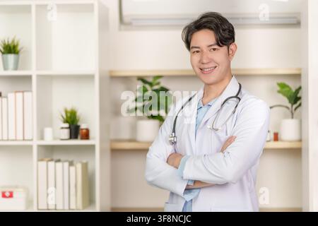 Asiatischer männlicher Arzt in weißem Kittel, der mit verschränkten Armen in der Klinik steht Stockfoto