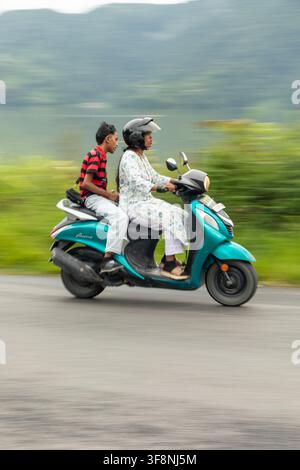Pokhara, Gandaki-Provinz, Nepal - 02. Juli 2026: Blick auf eine Frau, die einen türkisfarbenen Roller fährt, auf dem ein junger Mann mitfährt, entlang einer Straße mit einem verschwommenen Gr Stockfoto