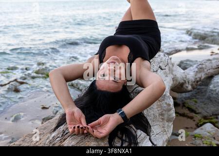 Selbstbewusste Frau in schwarzem Kleid, die auf Treibholz am Meer liegt und die Meeresküste mit Wellen und natürlicher Landschaft im Hintergrund genießt. Stockfoto