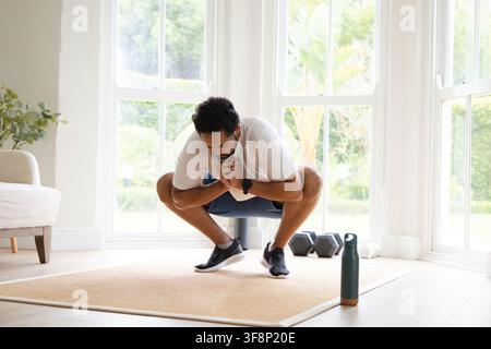 Mittelalter Afrikaner führt auf einer beigen Matte zu Hause in der Nähe von Hanteln und einer Wasserflasche einen tiefen Kniebeugen durch Stockfoto