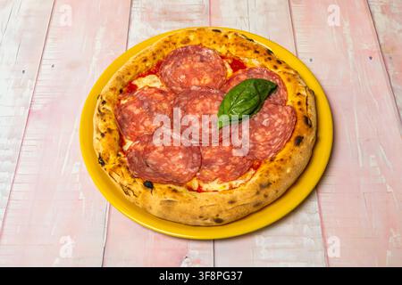 Holzofenpizza mit dicken Salamischeiben und Basilikumblatt, serviert auf einem hellgelben Teller. Stockfoto