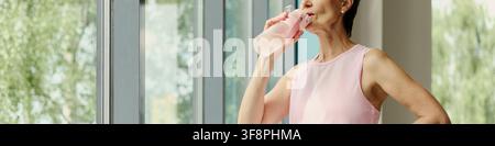 Reife kaukasische Frau trinkt Wasser aus der Flasche nach dem Training im Fitnessstudio, fördert einen gesunden Lebensstil. Bild geeignet für Kampagnen zur aktiven Alterung und Fitness-Marketing, mittlere Aufnahme Stockfoto