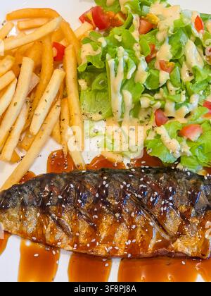 Ein gegrillter Makrele mit süßer Sojasauce glasierter, serviert mit Pommes frites und frischem Kopfsalat, der mit cremiger Sauce, Tomatenstücken und Sesam garniert ist Stockfoto