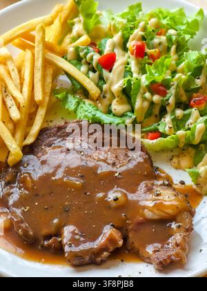 Ein gegrilltes Steak mit brauner Soße, serviert mit Pommes frites und frischem grünen Salat mit cremiger Dressing, warmes Wohlfühlessen auf weißem Teller, appetitlicher Din Stockfoto