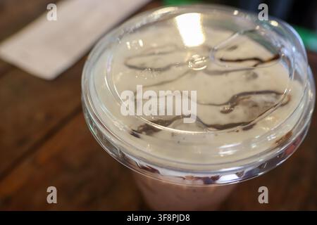 Ein gekühltes Schokoladengetränk mit Schlagsahne und Schokoladensoße in einem Plastikbecher auf einem Holztisch Stockfoto