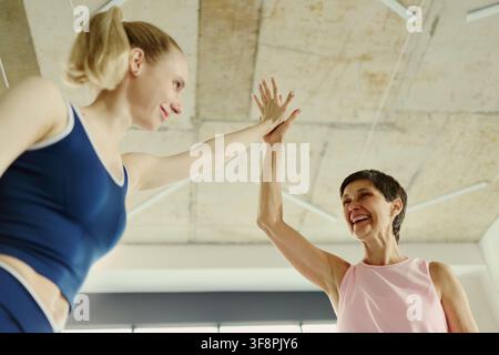 Junge kaukasische Frau und ältere kaukasische Frau geben sich im Fitnessstudio ein High Five und feiern den Fitness-Erfolg. Tiefohnaufnahme nützlich für Kampagnen zum aktiven Altern und intergenerationale Wohlbefindenskampagnen Stockfoto