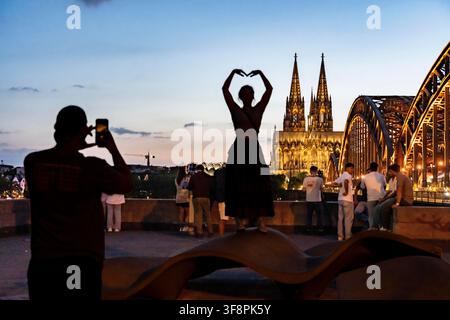 Blick auf den Kölner Dom vom Kennedy Ufer in Köln-Deutz; Passanten und Touristen, die nach Sonnenuntergang Fotos machen; Herzen, die sie mit ihren Haaren formen Stockfoto