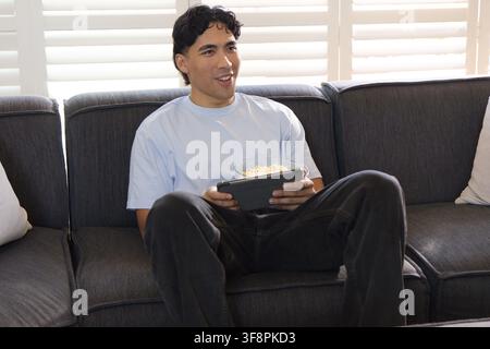 Asiatischer Mann sitzt auf dunklem grauem Sofa im Wohnzimmer, hält eine Popcorntschale und nutzt ein Tablet Stockfoto