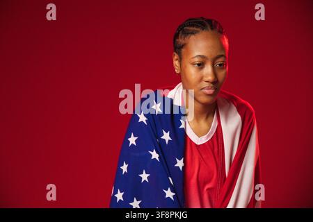 Afroamerikanerin posiert im Studio, wickelt sich in amerikanische Flagge, trägt rotes Hemd, Cornrows, Nieten Stockfoto