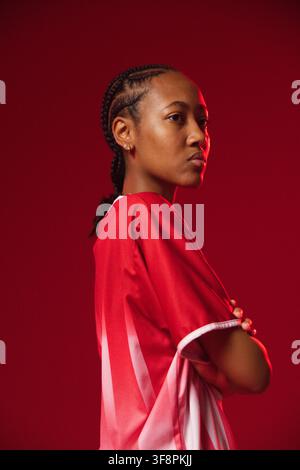 Afroamerikanische Frau im Profil im Studio posiert, trägt rotes-weißes Sporttrikot, Zöpfe, Kreolen Stockfoto