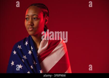 Afroamerikanerin posiert eingewickelt in US-Flagge, blickt nach links im Studio, geflochtene Haare, Kopierraum Stockfoto