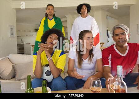 Verschiedene Freunde in Trikots, die zu Hause ein Fußballspiel anschauen, halten einen Fußball und Bierflaschen Stockfoto