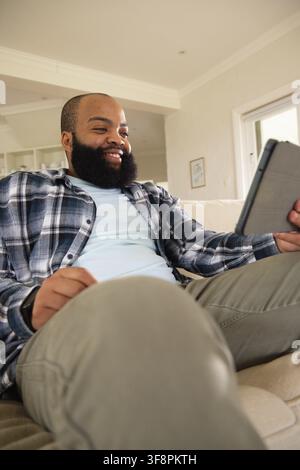 Afroamerikanischer Mann sitzt auf cremefarbenem Sofa im Wohnzimmer, hält ein Tablet und lächelt auf den Bildschirm Stockfoto