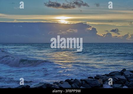 Dramatischer Sonnenuntergang über dem Ligurischen Meer, mit Wellen, die entlang einer felsigen Küste unter glühenden Wolken in Ligurien, Italien, brechen. Stockfoto
