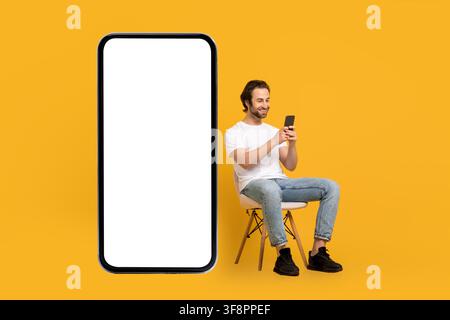 Glücklicher junger Mann, der ein Smartphone benutzt, sitzt auf einem Stuhl, gelber Hintergrund, Technologiekonzept Stockfoto