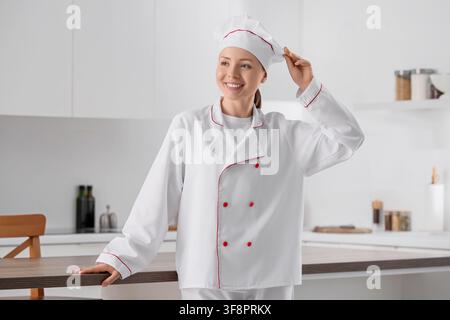 Lächelnde junge Chefköchin in einer weißen Uniform mit rotem Rand und Toque, steht in einer modernen Kücheneinrichtung, die kulinarische Fachkraft verkörpert Experti Stockfoto