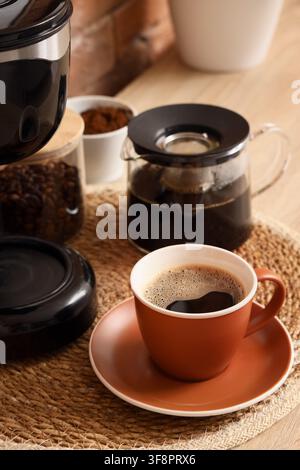 Frisch gebrühter schwarzer Kaffee in einer Terrakotta-Tasse mit Untertasse, umgeben von Kaffeebohnen und gemahlenem Kaffee Stockfoto