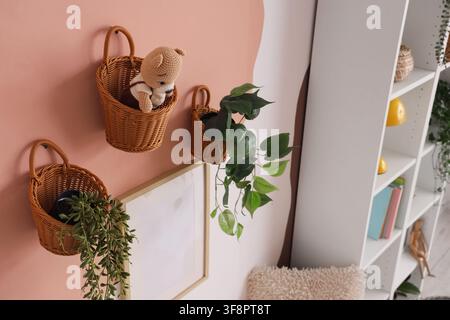 Weidenkörbe mit Pflanzen und ein Teddybär, der an einer pfirsichfarbenen und weiß gestrichenen Wand in einem gemütlichen Raum hängt, ein weißes Bücherregal mit Büchern und Deko-Artikel Stockfoto