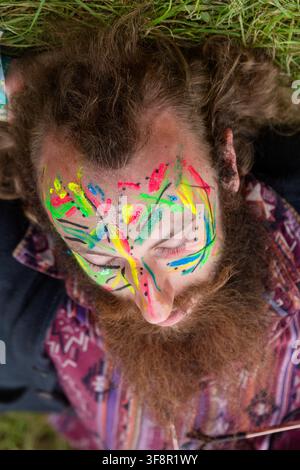 FESTIVALBESUCHER MIT NEON-GESICHTSBEMALUNG, REPORTAGE, GREEN MAN FESTIVAL 2016, BRECON, WALES, UK, 2016: Eine Nahaufnahme von oben zeigt einen bärtigen Mann mit aufwendiger, farbenfroher Neon-Gesichtsbemalung, der mit geschlossenen Augen im Gras liegt. Foto: Rob Watkins. INFO: Das Green Man Festival begann 2003 in Wales. Es findet jährlich im dritten Wochenende im August in den Brecon Beacons statt und feiert alternative Musik, Kunst, Literatur, Wissenschaft und familienfreundliche Kultur in einer wunderschönen Bergkulisse. Stockfoto