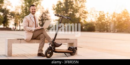 Lächelnder Geschäftsmann arbeitet an einem Laptop, während er auf einer Bank neben einem E-Scooter in einem Park bei Sonnenuntergang sitzt, ein E-Scooter ist daneben geparkt Stockfoto