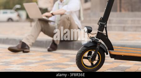 Elektro-Scooter, der auf einer Stadtstraße geparkt ist, mit einem Mann, der im Hintergrund an einem Laptop arbeitet, urbane Mobilität und Fernarbeit Stockfoto