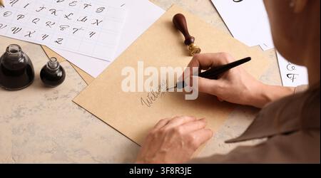 Person schreibt 'willkommen' in Kalligraphie mit einer Füllfeder auf braunem Papier, umgeben von Tintenflaschen und Alphabet-Übungsblättern Stockfoto