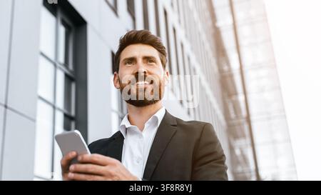 Lächelnder junger kaukasischer Mann, der im Freien ein Smartphone in einem Business-Anzug hält, Business-Konzept Stockfoto