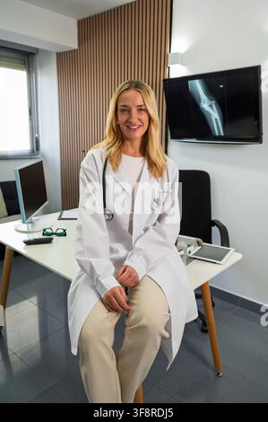 Ärztin sitzt auf einem Schreibtisch in einer medizinischen Klinik und lächelt in die Kamera Stockfoto
